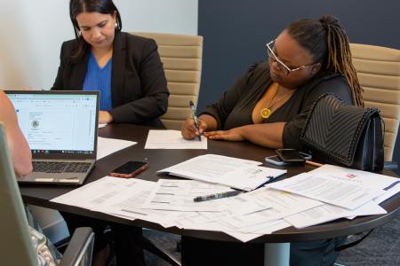 Two women are seated at a conference table, engaged in paperwork. The table is covered with various documents, pens, and a laptop showing an email. One woman, wearing a blue shirt and black blazer, is concentrating on reading a paper. The other woman, wearing glasses and a black top with a gold necklace, is writing on a document. A black handbag and two smartphones are on the table near her. The atmosphere appears professional and focused, likely a business or legal setting.